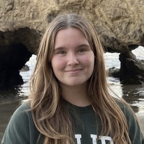 Evelina, smiling, on a beach with rock arches and waves behind her